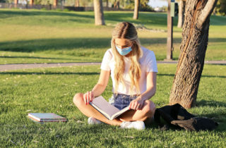 年轻的蒙面女大学生在户外阅读课本。