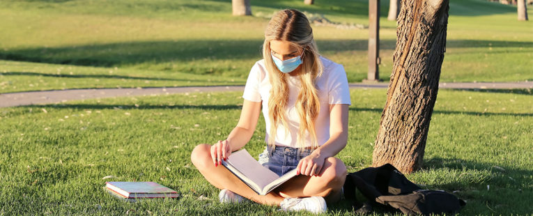 Young masked woman college student reading textbook outdoors.