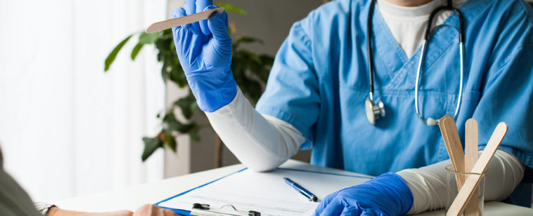 医生在办公桌前戴着口罩和手套，带压舌板;病人的手在前景可见