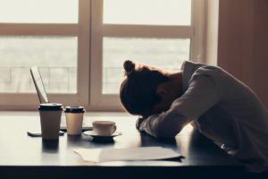 visibily fatigued woman slumped over her desk surrounded by cups of coffee