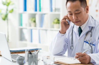 Male Asian doctor at desk speaking on phone with notepad and laptop in front of him