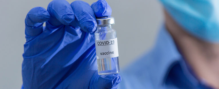 背景中穿着蓝领衬衫和蓝色外科口罩的医务人员戴着蓝色乳胶手套，用两根手指拿着前景中的疫苗瓶