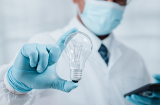 Doctor in surgical mask holds light bulb in foreground with blue gloved hand